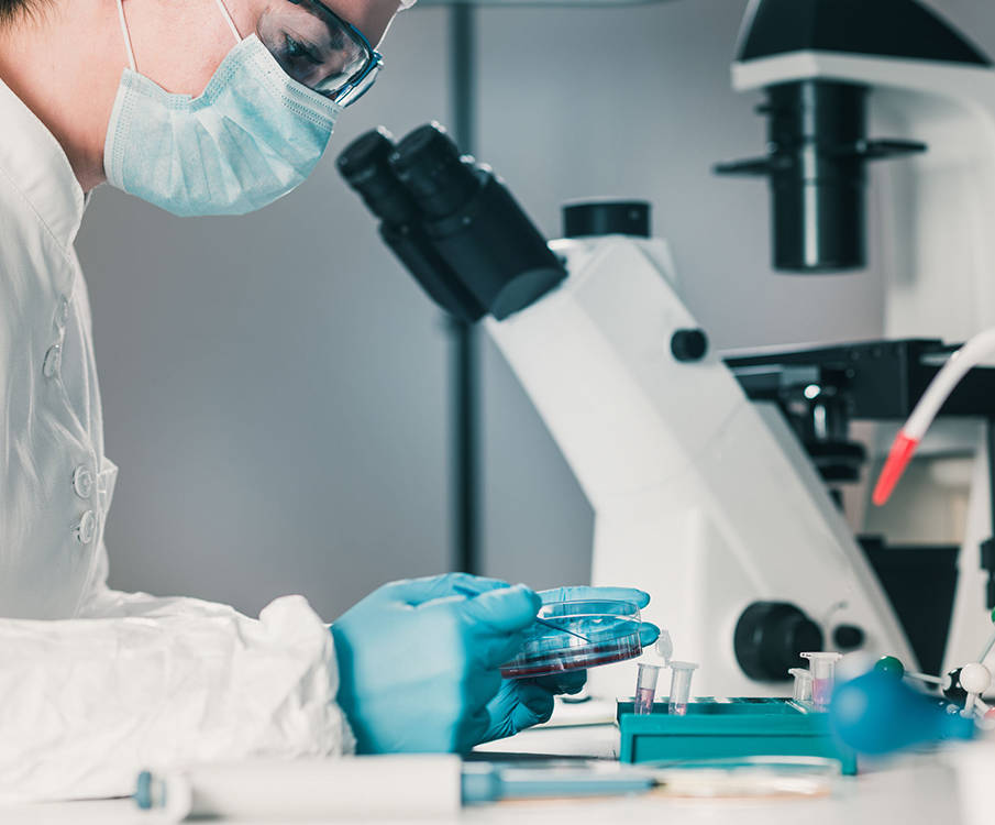 A laboratory technician with glasses and a mask examines the analysis in her hand and has a microscope on the table in the background