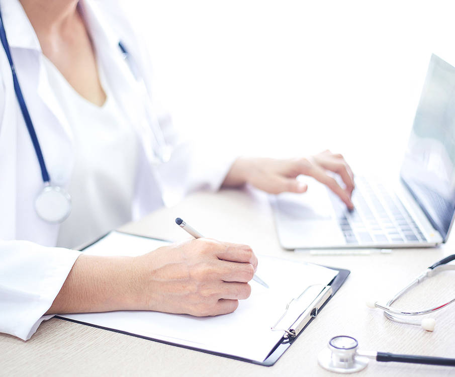 A female doctor with a stethoscope on a laptop and writes on paper with a pen