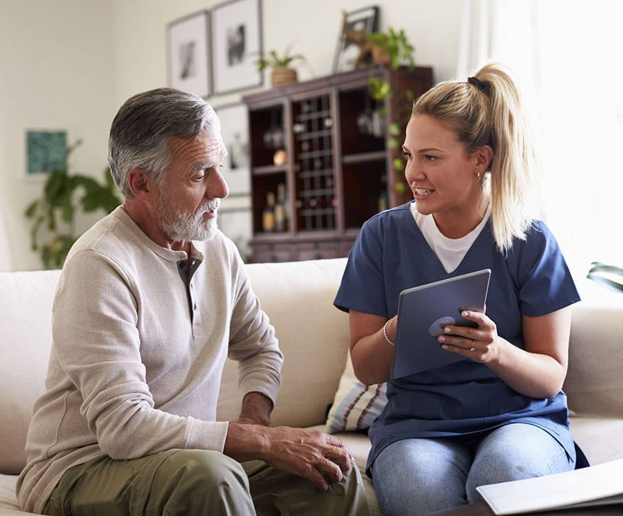 The nurse sits on the couch with the older man and shows the tablet