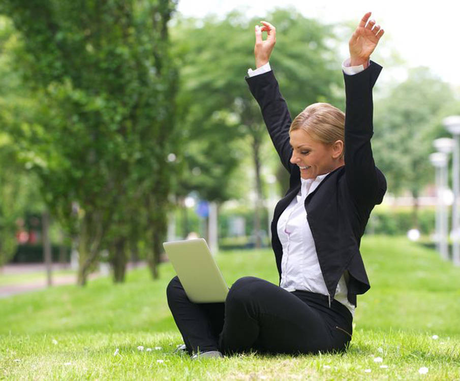 A happy woman sits on the grass with a laptop on her knees